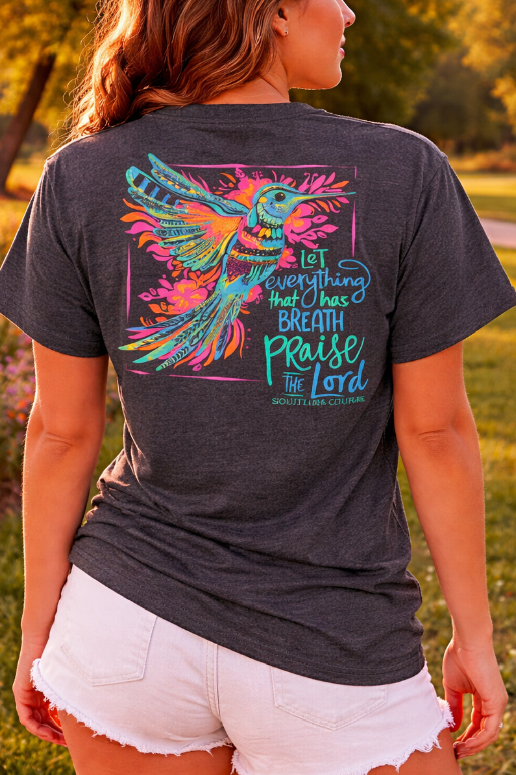 A woman stands outdoors in a park at sunset wearing a dark charcoal gray t-shirt with a colorful graphic of a bird and the text 'Let everything that breaths praise to Lord' on the back, paired with white denim shorts.