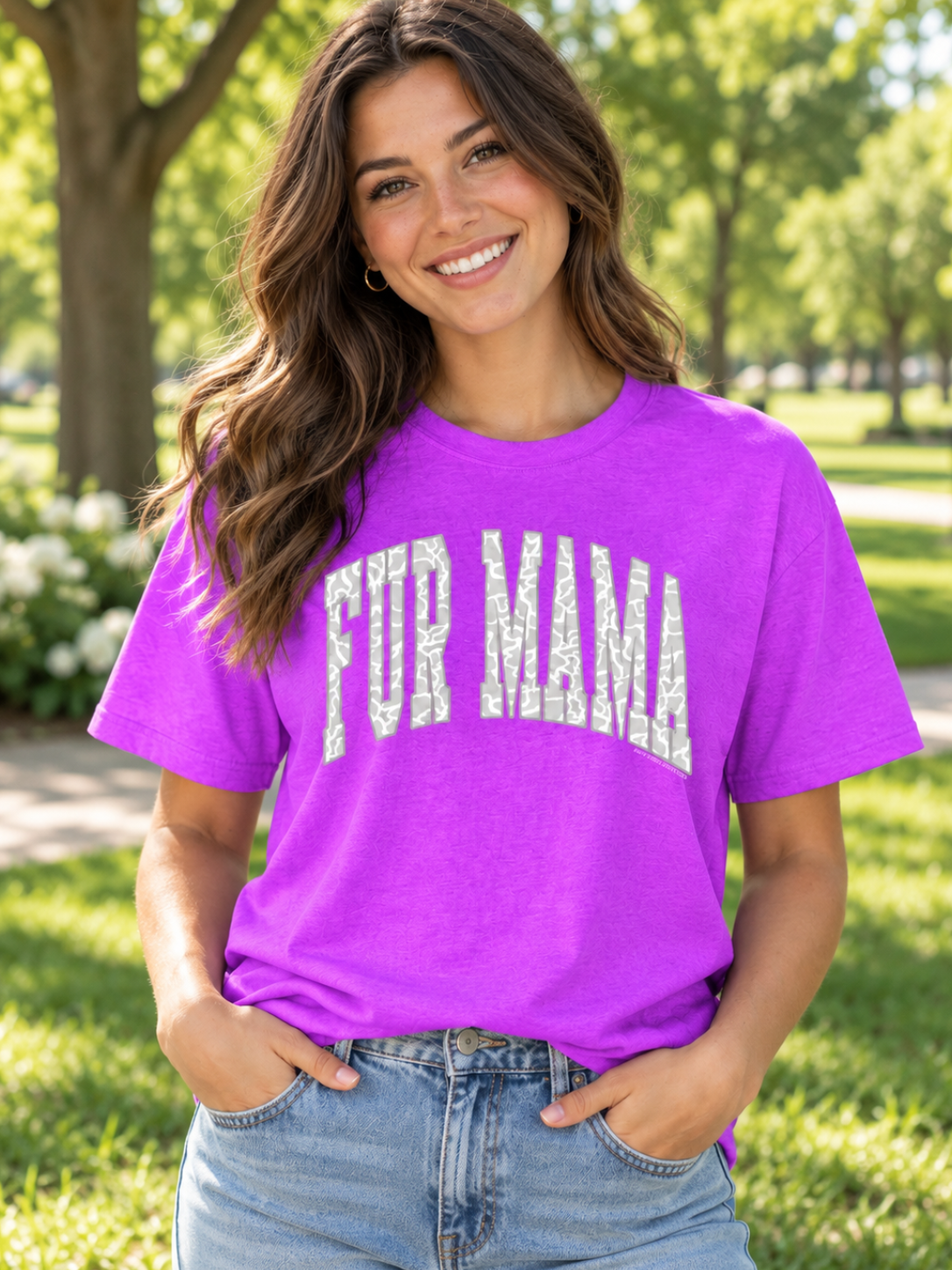 A woman smiling outdoors in a bright purple short-sleeve t-shirt with 'FUR MAMA' printed across the front in distressed white letters, paired with light blue jeans.