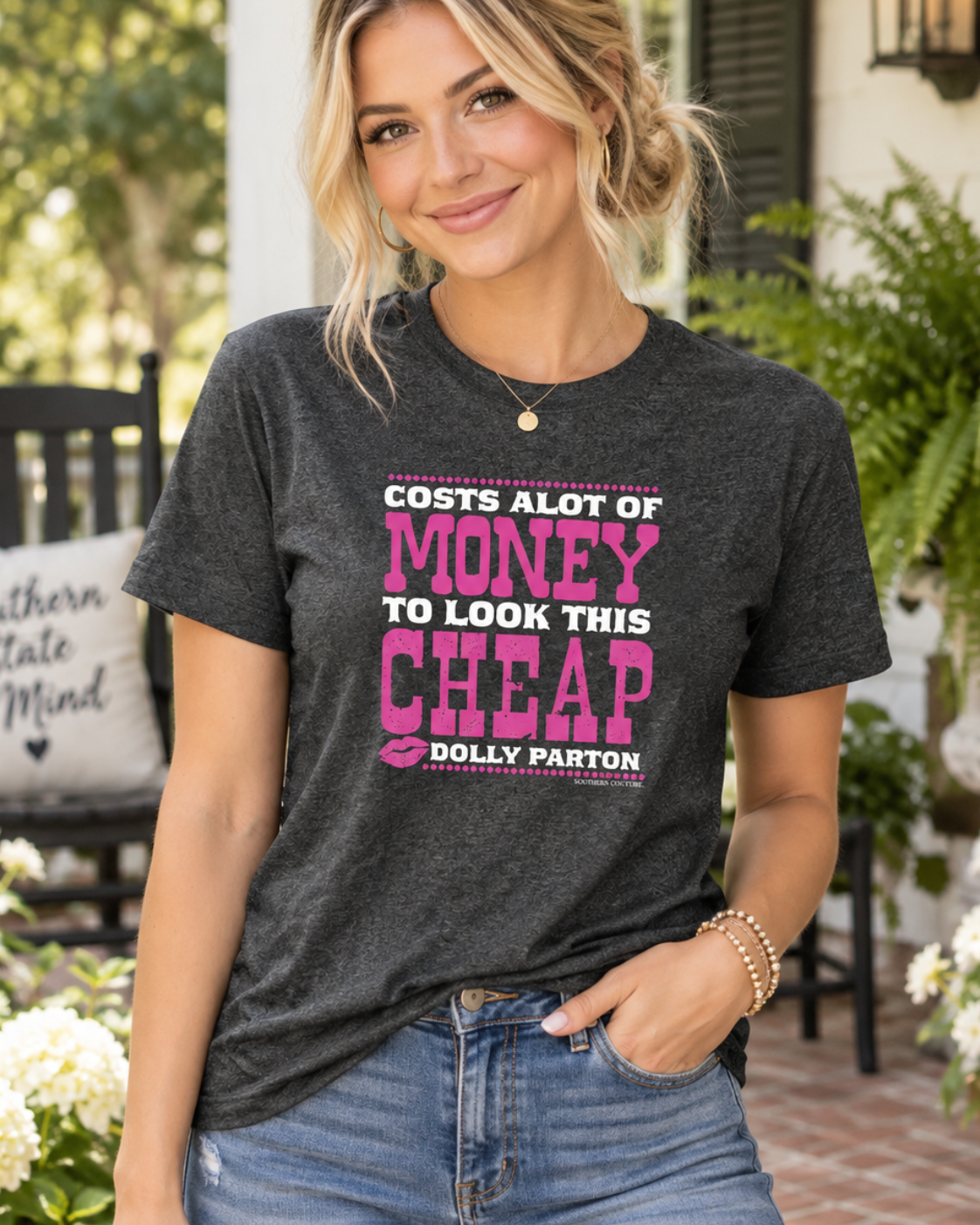A woman wearing a dark heather gray graphic t-shirt with pink and white text that reads 'COSTS ALOT OF MONEY TO LOOK THIS CHEAP DOLLY PARTON' paired with blue jeans, standing on a brick porch with plants and a chair in the background.