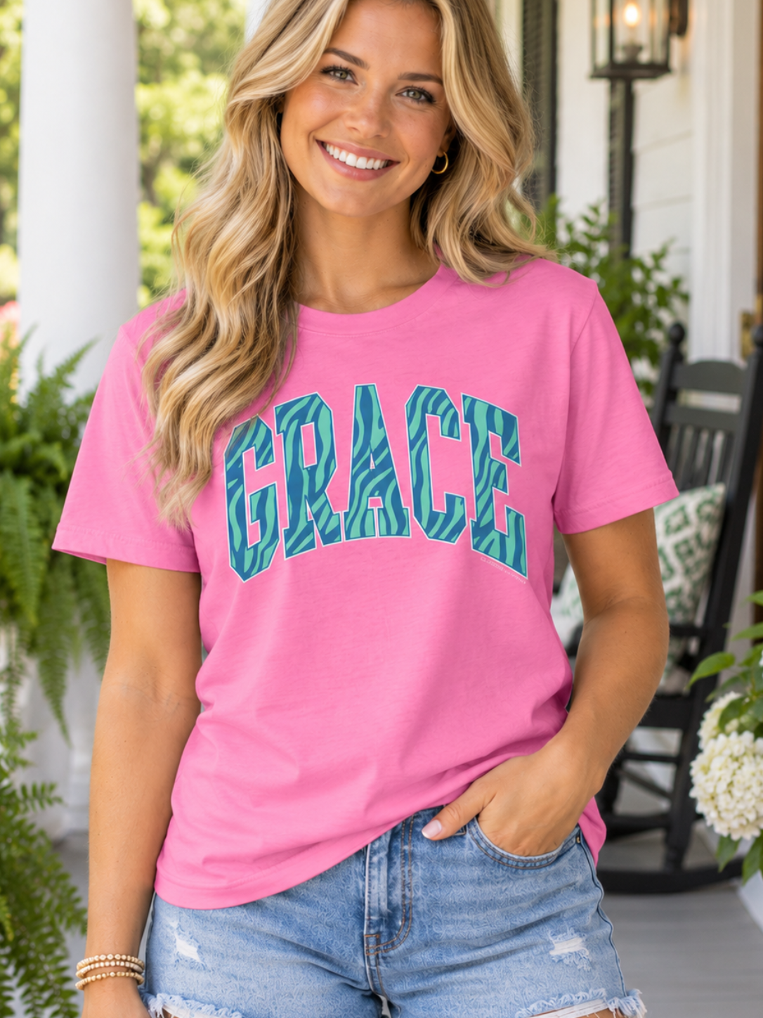 A woman stands on a porch wearing a bright pink short-sleeve T-shirt with the word 'GRACE' printed across the chest in teal patterned letters, paired with light blue denim shorts.