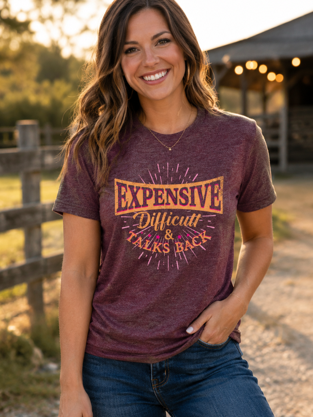 A woman wearing a heathered maroon graphic t-shirt with the text 'EXPENSIVE Difficult & TALKS BACK' in orange and yellow, paired with blue jeans, standing outdoors in front of a rustic barn at sunset.